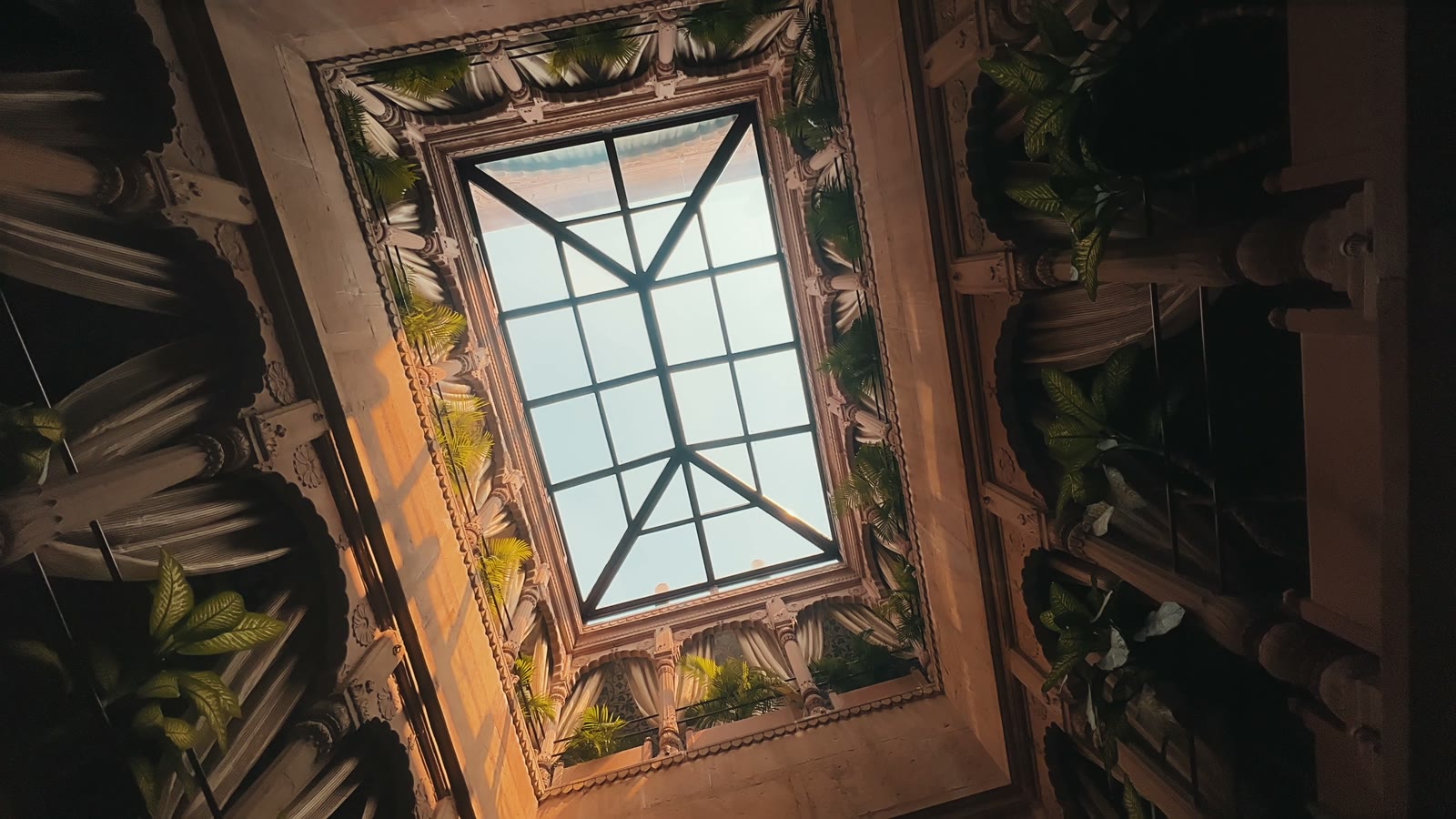 A glass skylight seen from below, framed by carved stonework and palms